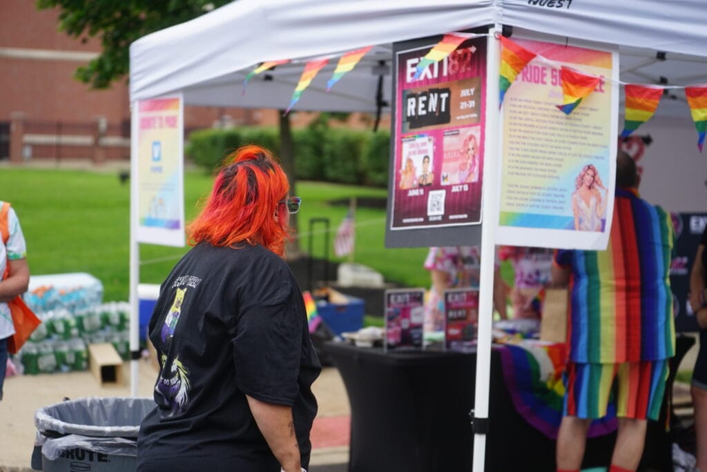 Attendee with bright orange hair browses a Pride booth decorated with rainbow flags and event posters at Toms River Pride.
