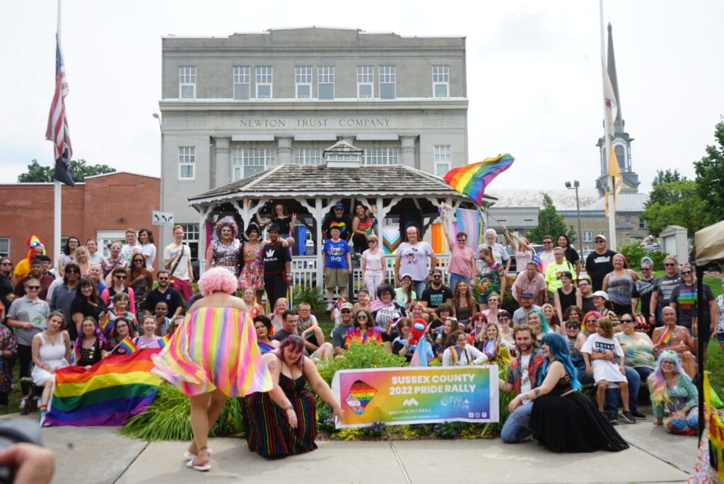 Large group gathered with rainbow and trans pride flags in front of a gazebo during Sussex County Pride Rally 2022 in Newton, New Jersey.