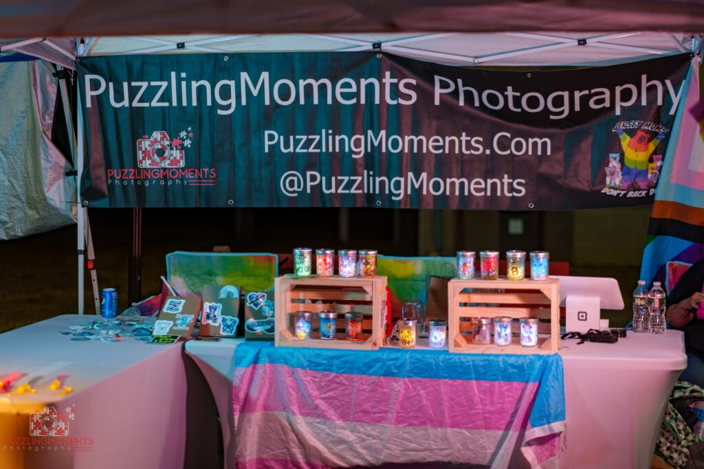 Vendor booth at Lunar Faire Pride Night Market featuring handmade items, glowing jars, and a transgender pride flag draped across the table under a “Puzzling Moments Photography” banner.