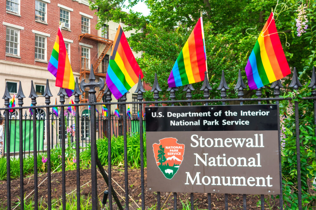Rainbow Pride flags line a fence outside the Stonewall National Monument, with a National Park Service sign visible in the foreground.